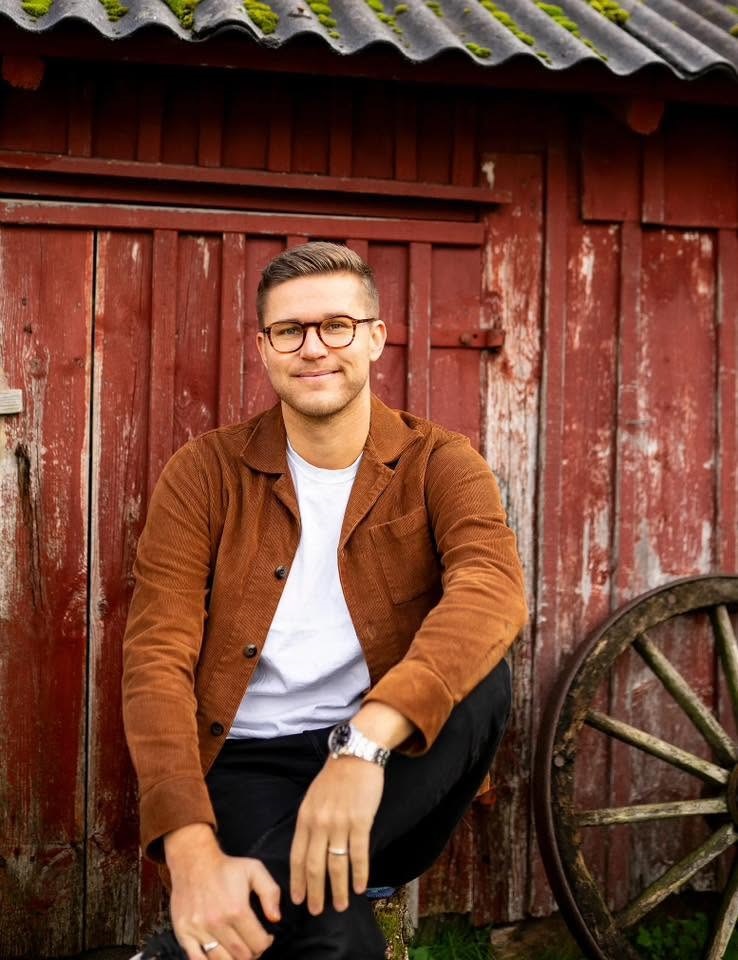 Leende man i brun jacka och glasögon sitter mot röd trÀvÀgg med gammalt hjul i bakgrunden.